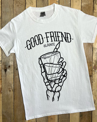 White t-shirt with black screen printed design of a skeleton hand holding a glass and 'GOOD FRIEND SLAINTE' text on a wooden background